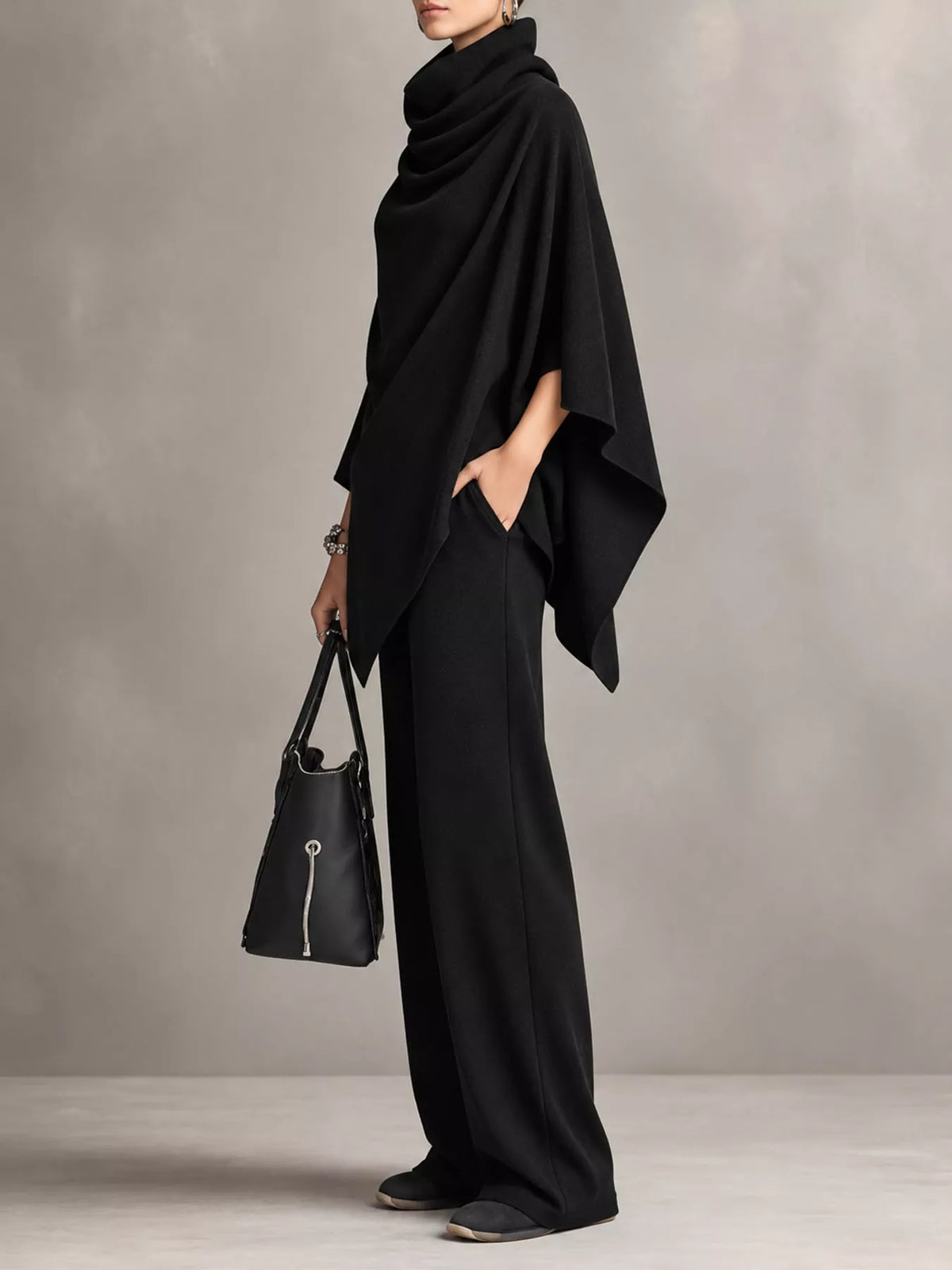 A model wearing a black draped poncho-style top, black wide-leg trousers, and holding a black leather handbag with silver hardware, set against a textured gray studio background.