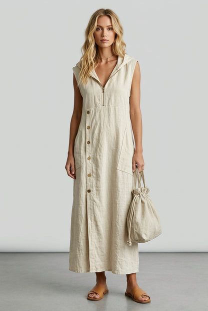 A woman wearing a sleeveless, off-white linen maxi dress with a front zipper, button-down front, and side pockets, holding a matching fabric bag in an outdoor garden setting.