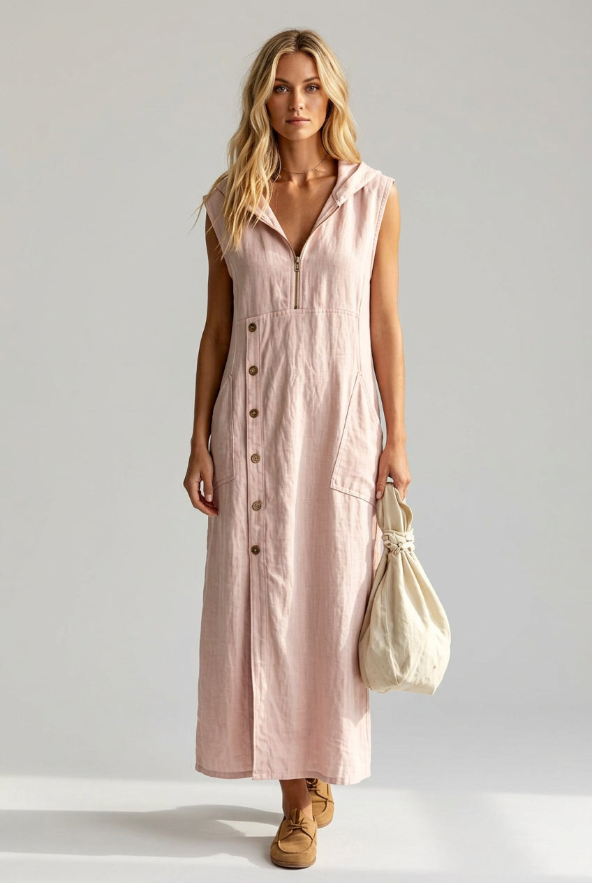 A woman wearing a sleeveless, light pink button-front midi dress with a collar and front pockets, standing outdoors near a white picket fence and greenery while holding a cream-colored drawstring bag.
