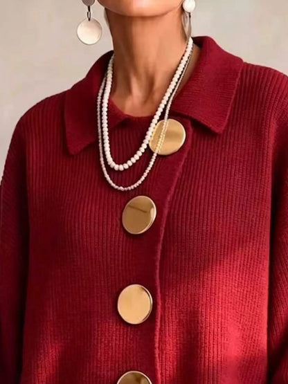 A close-up of a person wearing a deep red ribbed knit cardigan with oversized round gold buttons, layered with double-strand pearl necklaces and white disc earrings.