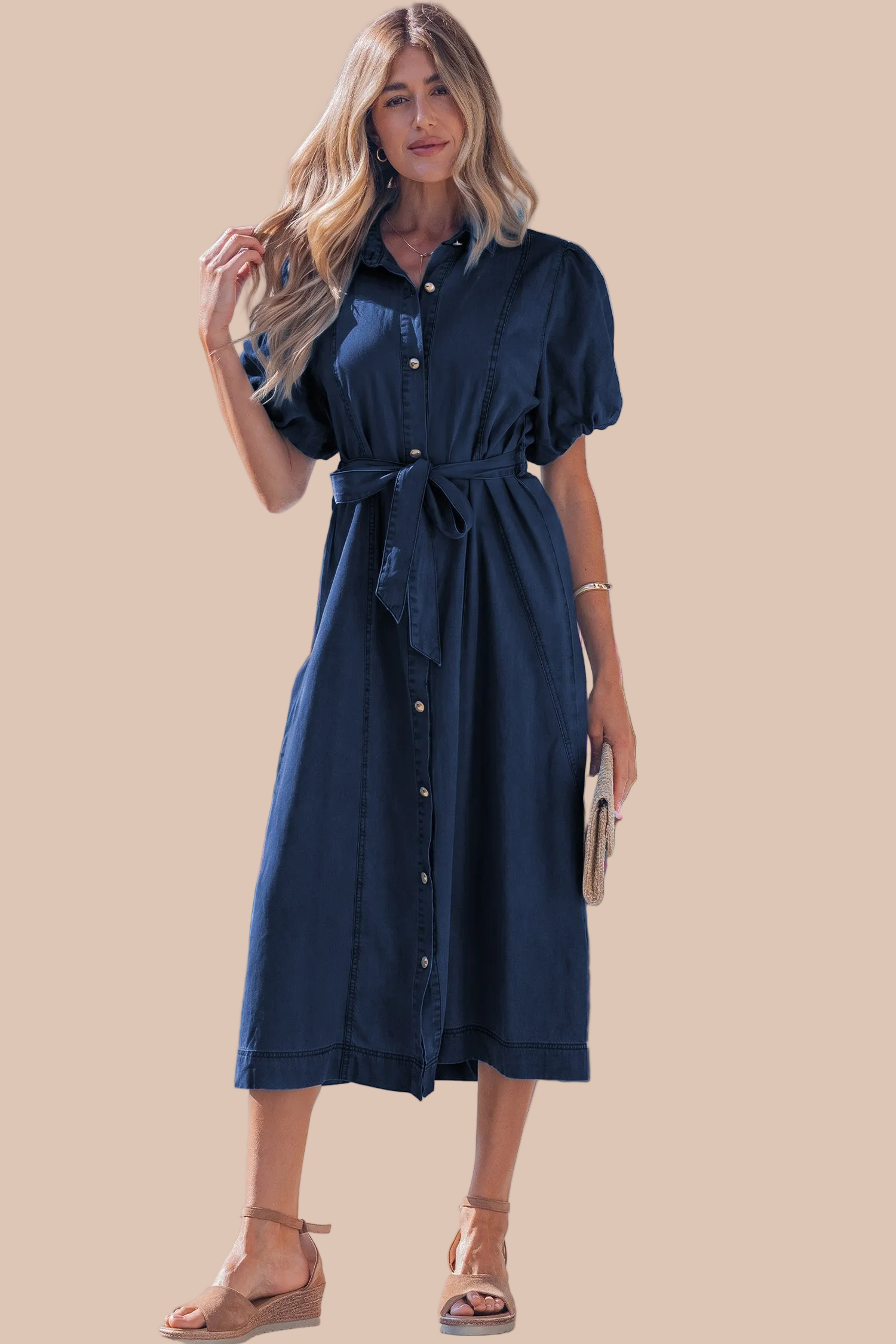 A woman modeling a dark navy blue midi shirt dress with short puff sleeves, a button-down front, and a matching fabric tie belt, set against a solid beige background.