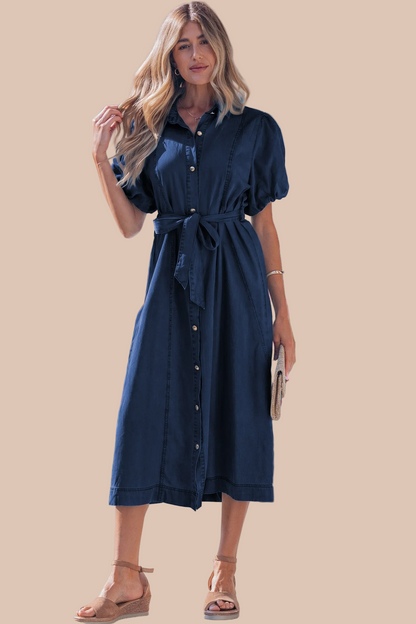 A woman modeling a dark navy blue midi shirt dress with short puff sleeves, a button-down front, and a matching fabric tie belt, set against a solid beige background.