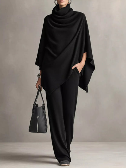 A model wearing a black draped poncho top and matching wide-leg trousers, holding a black leather handbag against a neutral gray studio background.