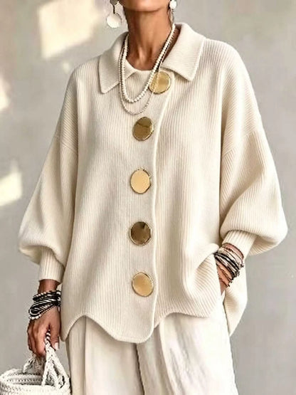 A person wearing an oversized cream ribbed knit cardigan with large round gold-tone buttons, paired with matching cream pants and layered pearl necklaces, against a softly lit neutral background.