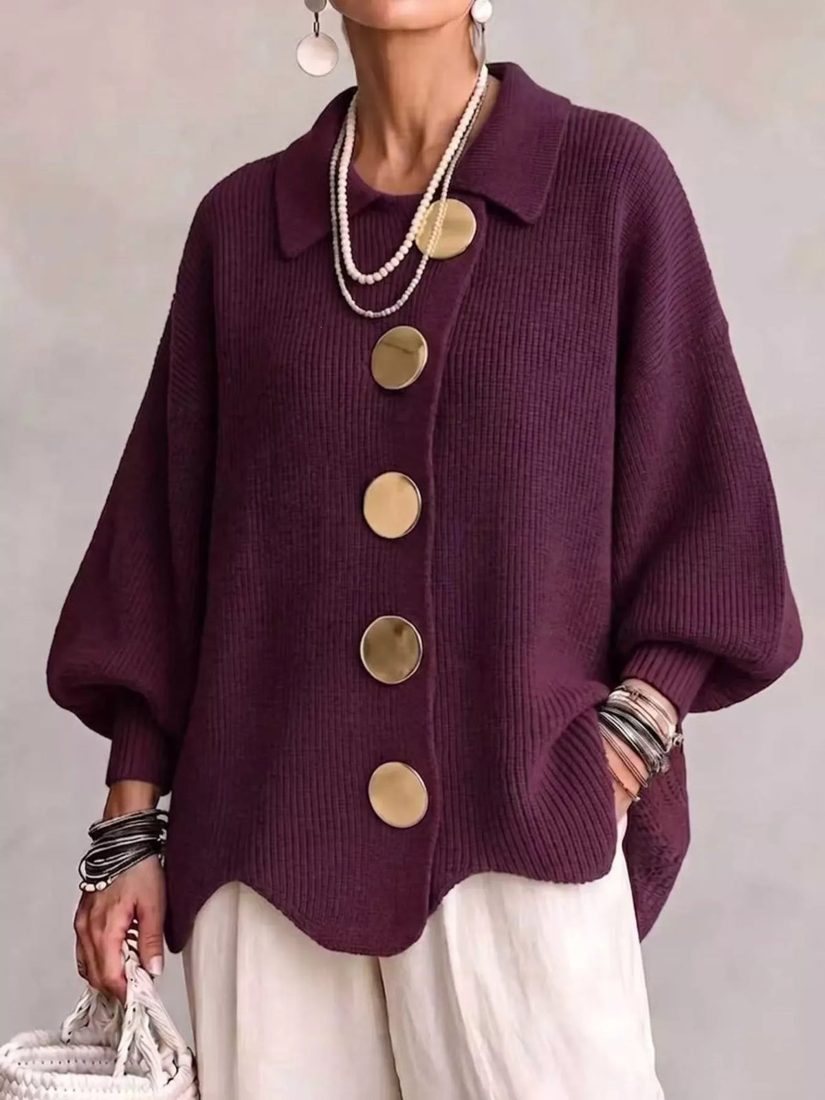 A person wearing a deep purple ribbed knit cardigan with oversized round gold-tone buttons, paired with cream pants and layered necklaces, against a neutral gray background.