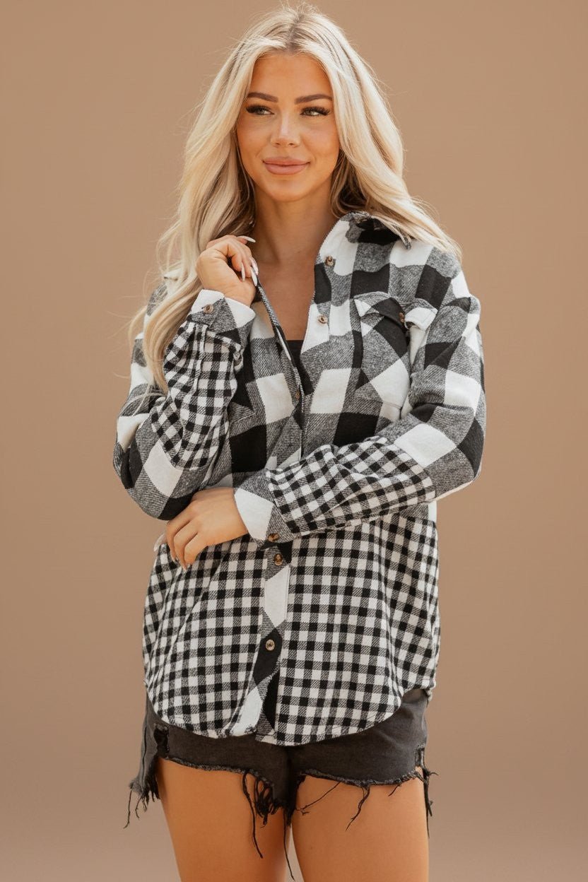 A woman wearing a black and white buffalo plaid button-up flannel shirt and dark denim shorts, standing outdoors in a sunny park setting with green grass and trees in the background.