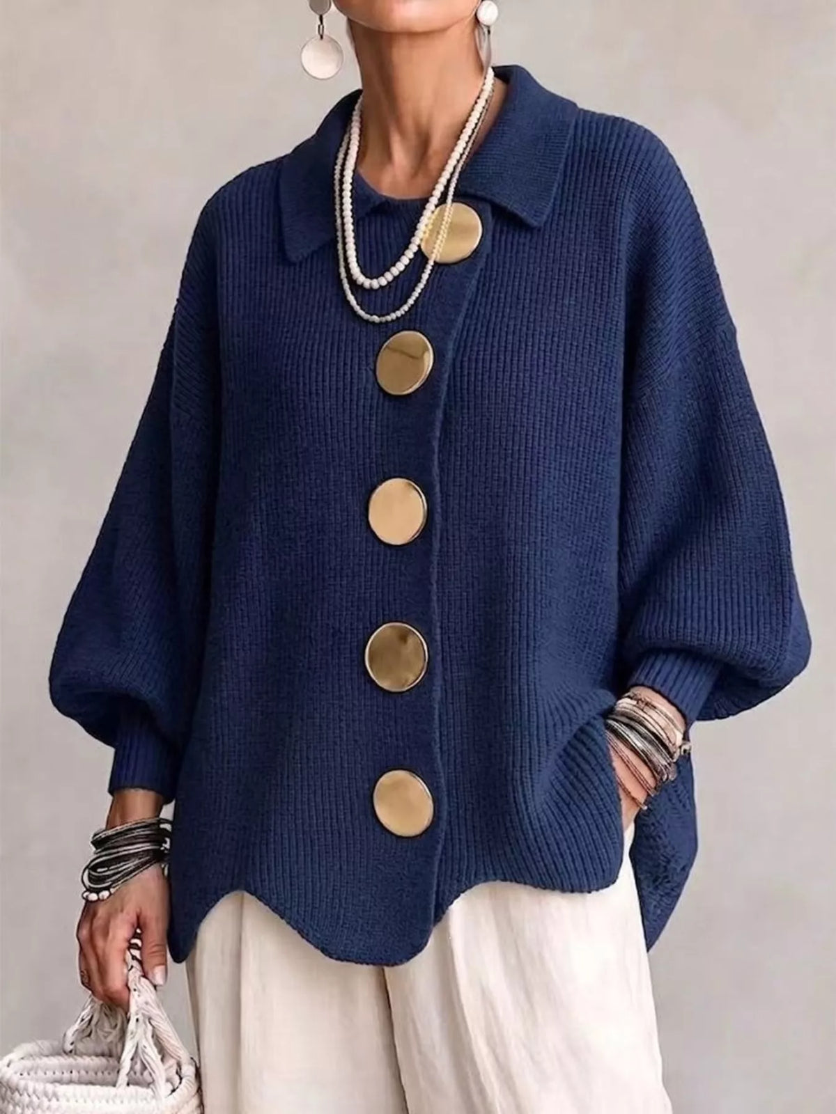 A woman wearing a navy-blue knit cardigan with oversized gold buttons, paired with cream pants, layered pearl necklaces, bracelets, and earrings.