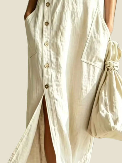 A person wearing a long cream linen shirt dress with a button-down front and side slit, holding a matching cream fabric bag outdoors.