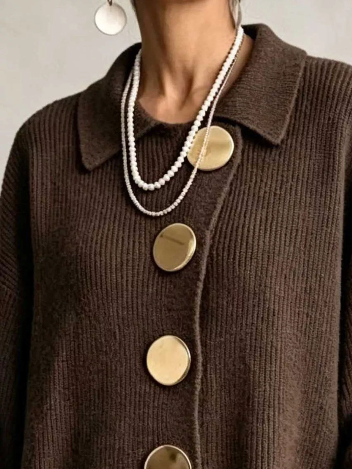 A close-up of a person wearing a dark-brown ribbed-knit cardigan with oversized, round gold buttons and layered white-pearl necklaces.