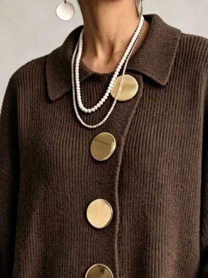 A close-up of a person wearing a dark-brown ribbed-knit cardigan with oversized, round gold buttons and layered white-pearl necklaces.