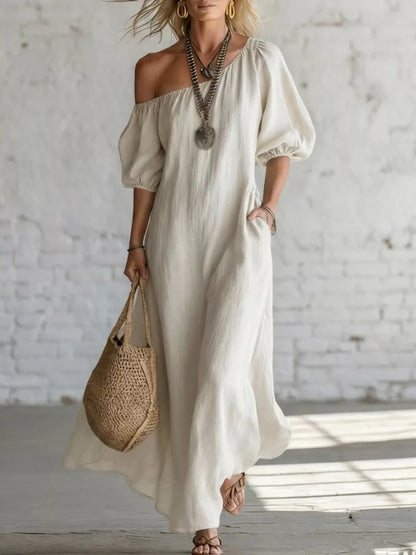 A woman wearing a flowing off-the-shoulder maxi dress in a light cream or off-white color, styled with layered silver necklaces, gold hoop earrings, a woven straw tote bag, and strappy sandals, standing against a white brick wall.