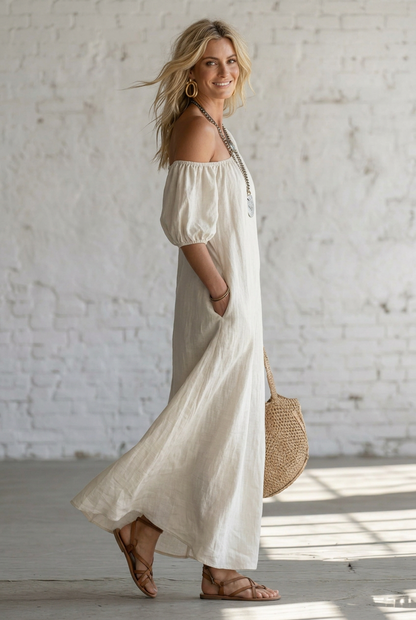 A woman wearing a flowing off-the-shoulder maxi dress in a light cream or off-white color, styled with layered silver necklaces, gold hoop earrings, a woven straw tote bag, and strappy sandals, standing against a white brick wall.