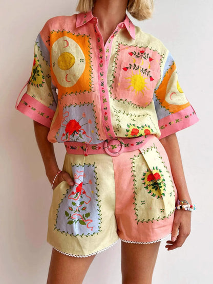A person wearing a pastel patchwork short-sleeve button-up shirt and matching shorts with embroidered floral and sun motifs, pink trim, and a pink belt against a plain light background.