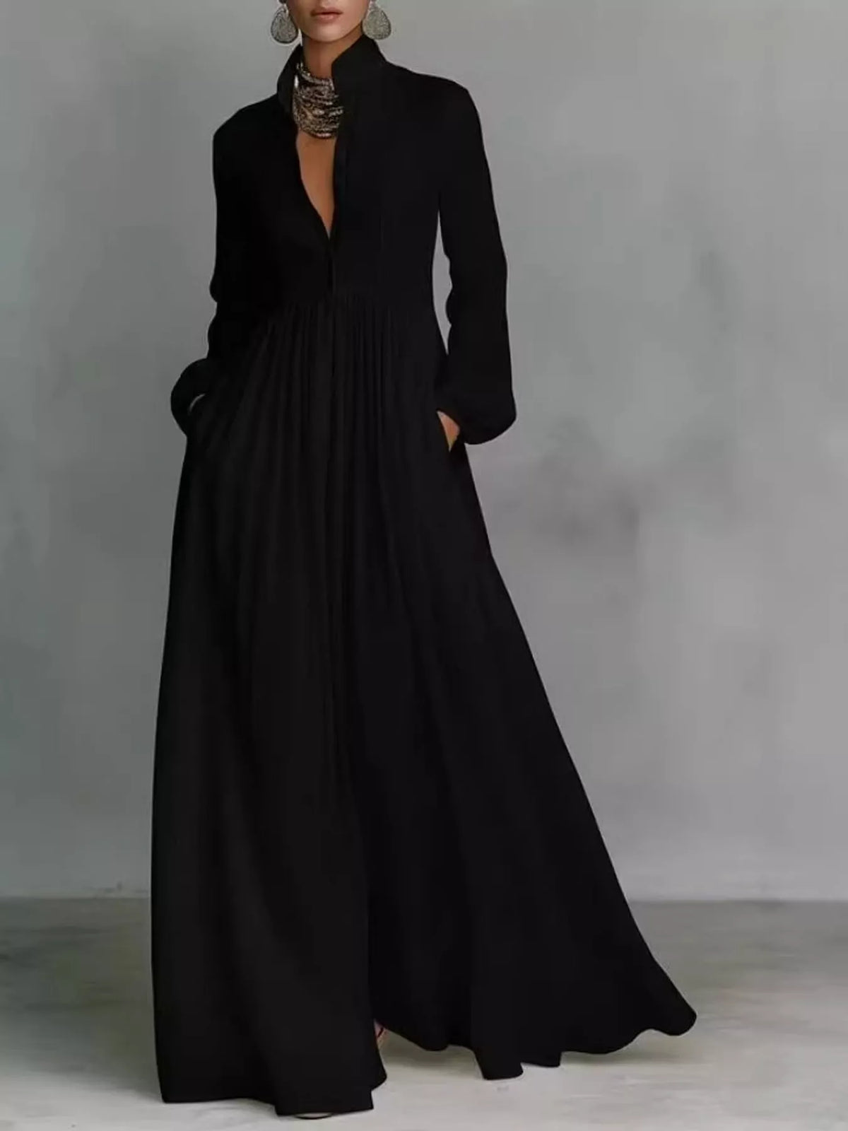 A woman modeling a long black maxi dress with a deep V-neckline, long sleeves, and side pockets, standing against a textured gray studio backdrop.