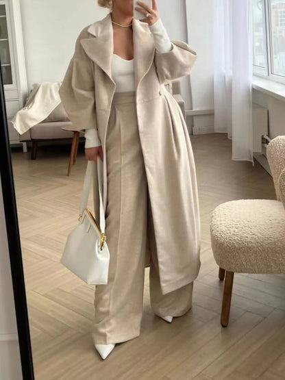 Person wearing a beige trench coat and wide-leg pants in a room with wooden flooring.