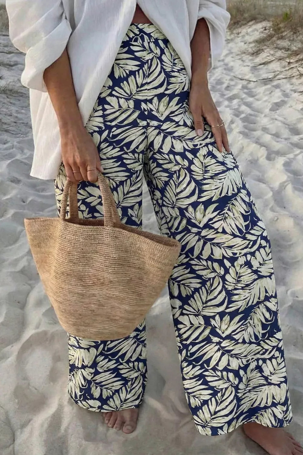 Person wearing blue and white leaf-patterned pants on a sandy beach, holding a woven bag.