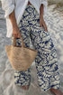 Person wearing blue and white leaf-patterned pants on a sandy beach, holding a woven bag.
