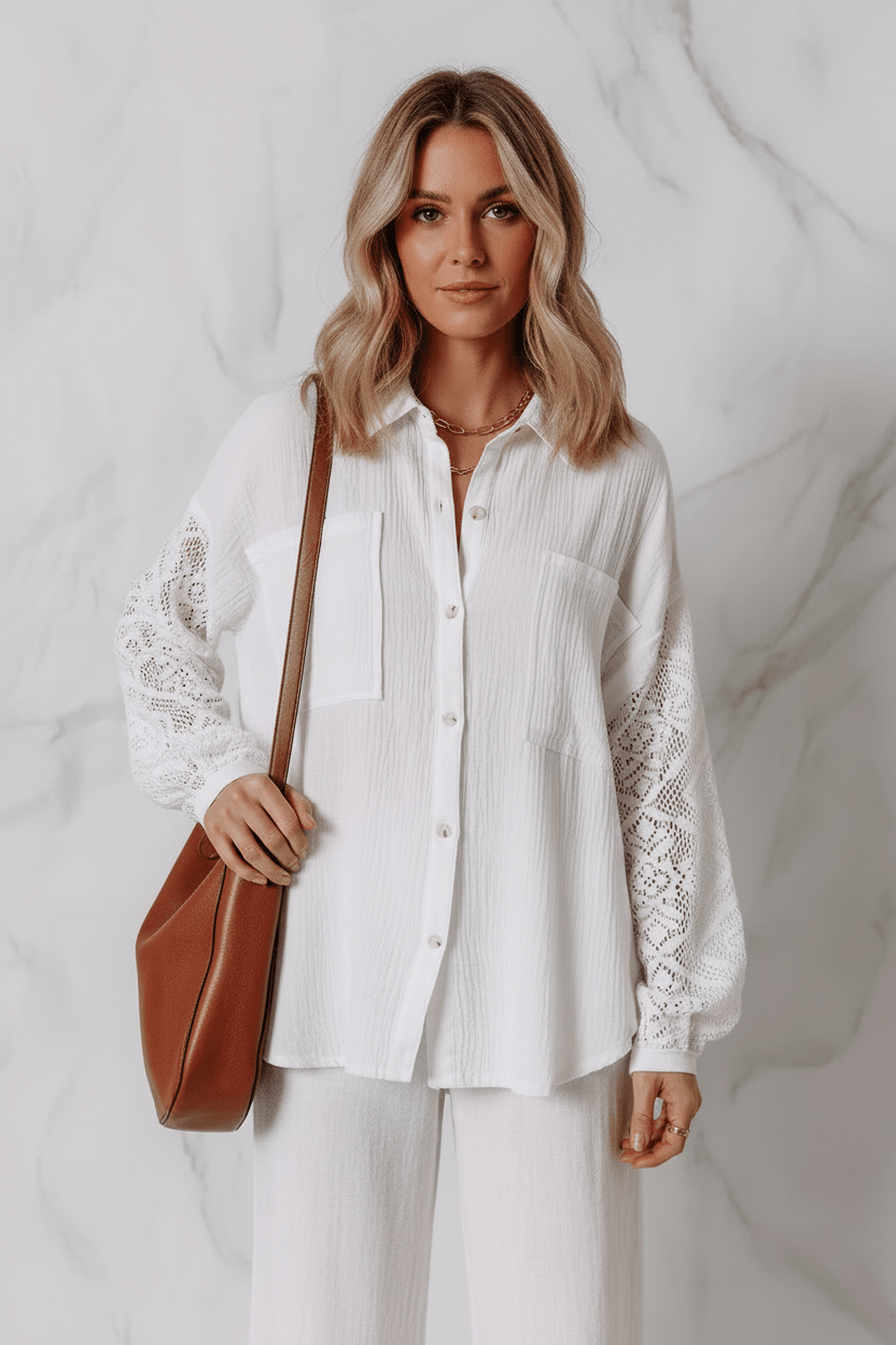 A woman wearing a white long-sleeve button-up blouse with lace sleeves and white pants, holding a brown leather shoulder bag against a light gray marble wall.