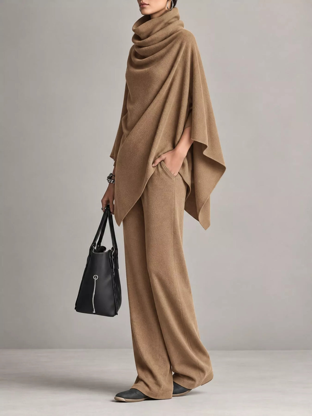 A model wearing a camel brown knit poncho and matching wide-leg trousers, holding a black leather handbag against a neutral gray studio background.