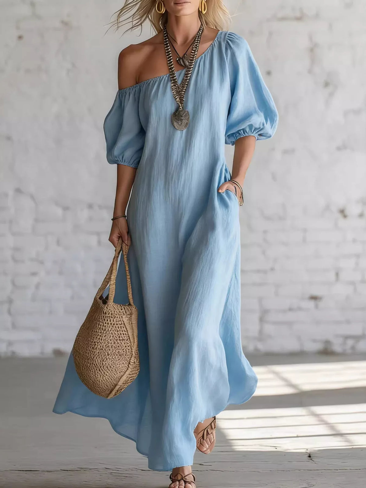 A woman wearing a light blue off-the-shoulder maxi dress with puff sleeves and side pockets, holding a woven straw tote bag against a white brick wall background.