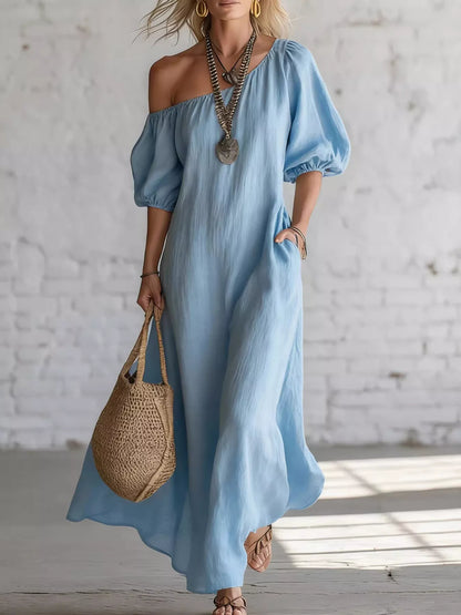 A woman wearing a light blue off-the-shoulder maxi dress with puff sleeves and side pockets, holding a woven straw tote bag against a white brick wall background.