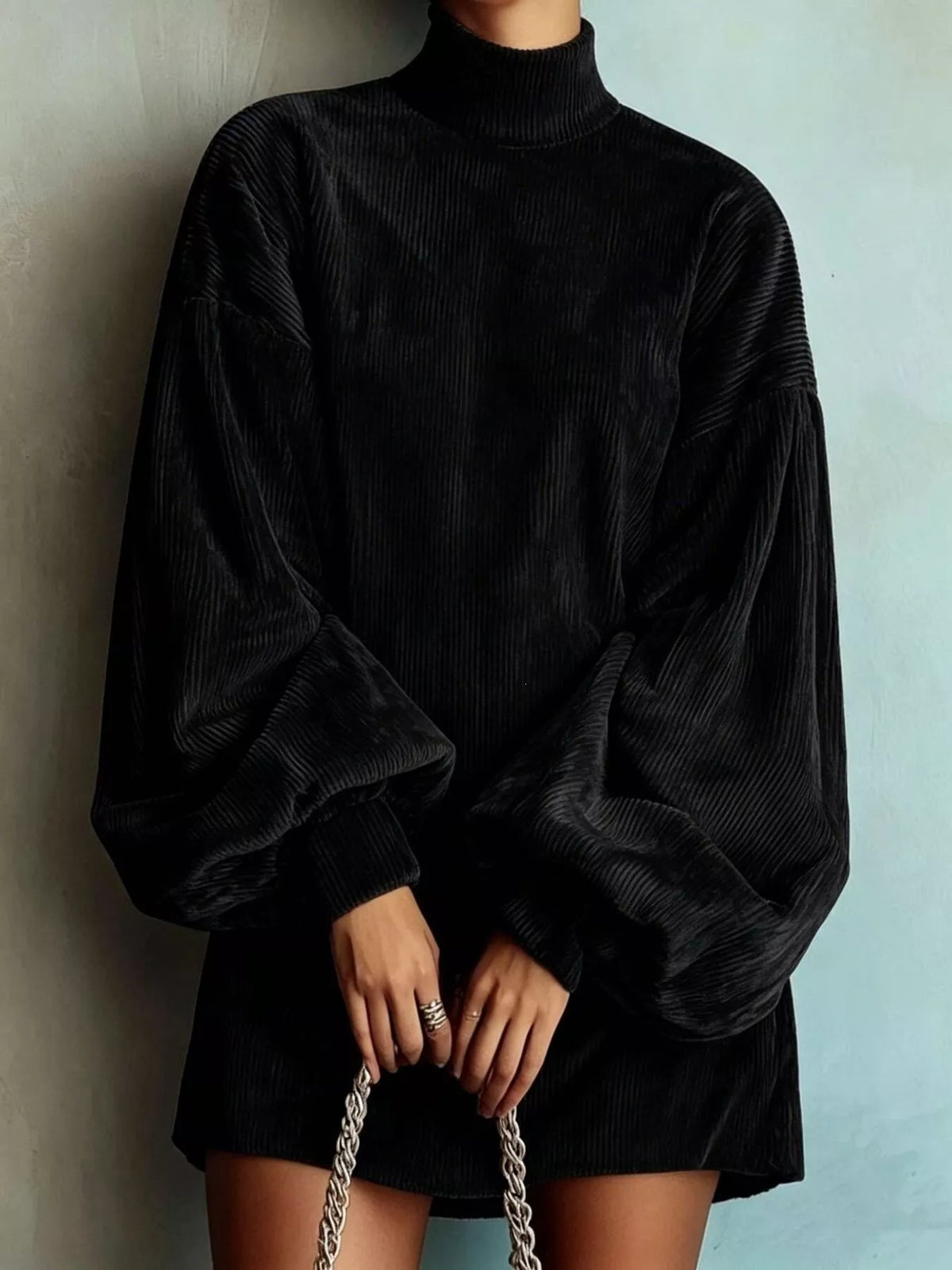 A person wearing an oversized black corduroy mock-neck top with dramatic balloon sleeves, paired with a matching black corduroy mini skirt, holding a silver chain bag against a textured light blue-gray wall.