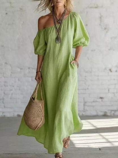 Woman wearing a light green off-shoulder dress holding a woven bag against a white brick wall.