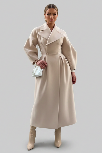 A woman wearing a long, double-breasted beige coat with wide lapels and voluminous sleeves, styled with a pearl-embellished white handbag and bracelet against a solid gray background.