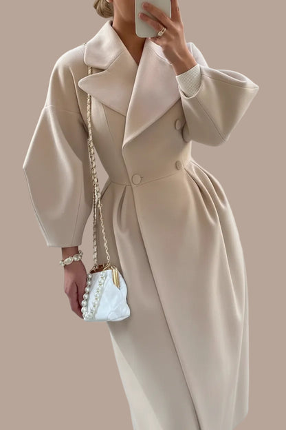 Person wearing a long beige coat holding a white handbag with pearls on a paved street.