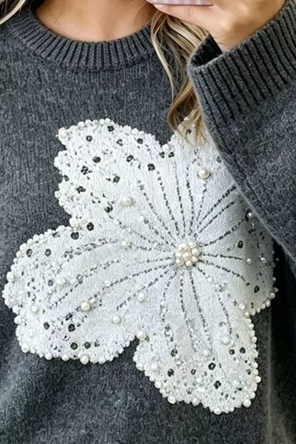 A close-up of a dark gray knit sweater featuring a large, detailed white floral applique on the front. The floral design is embellished with white pearls, clear rhinestones, and silver beadwork, with a model&