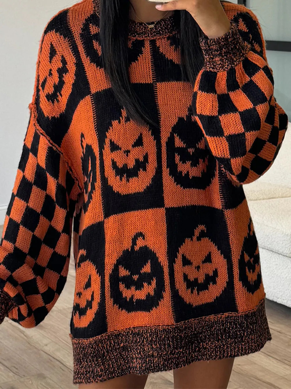 A person wearing an oversized knit sweater with a Halloween theme, featuring orange and black checkerboard patterns and large pumpkin faces.