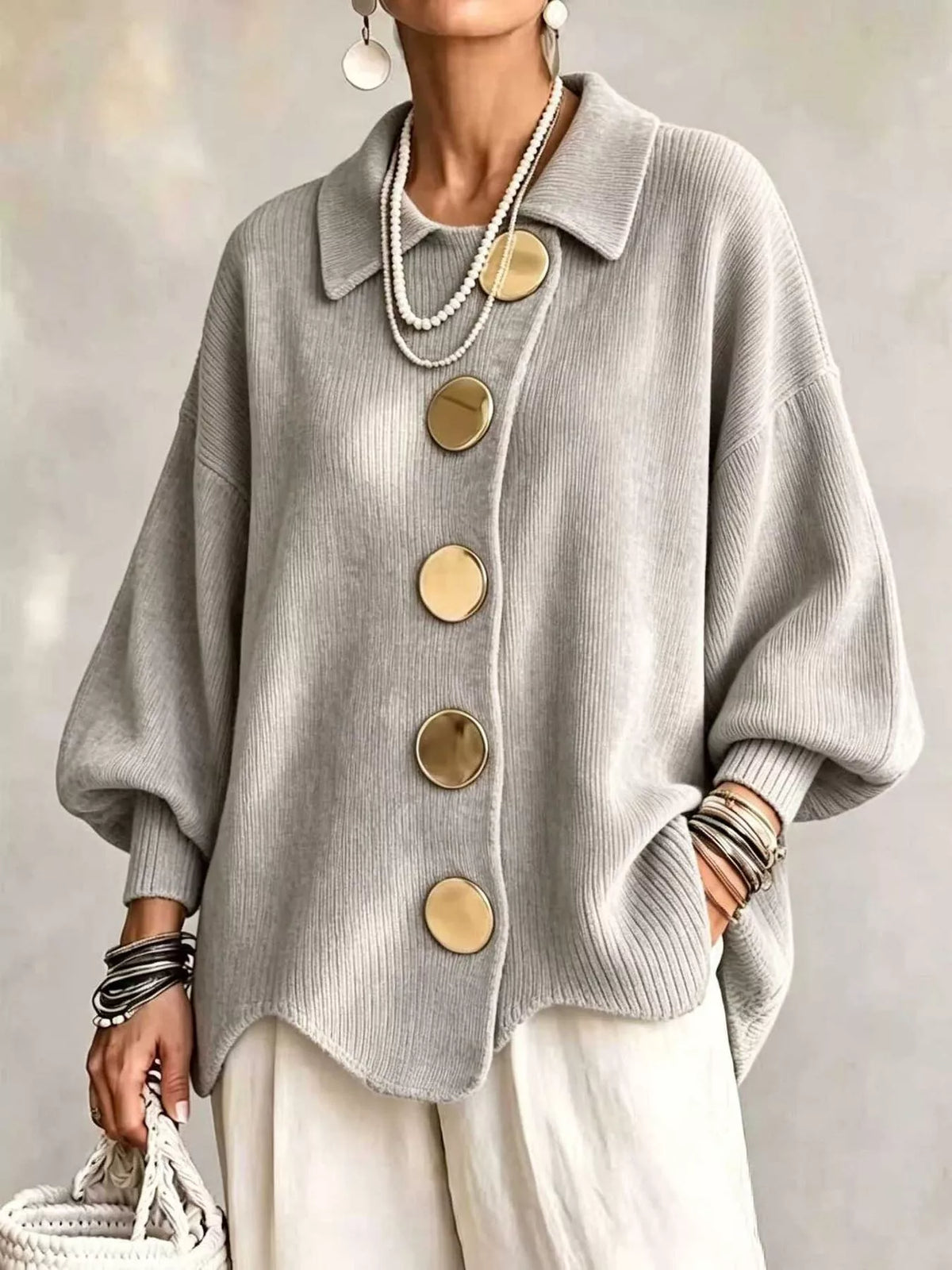 A woman wearing a light gray ribbed knit cardigan with oversized round gold buttons, styled with cream pants, layered pearl necklaces, bracelets, and earrings.