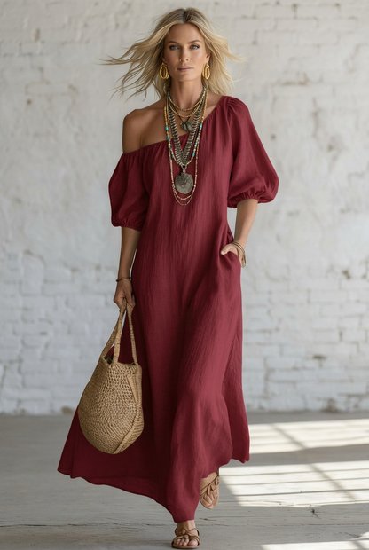 A woman wearing a flowing off-the-shoulder maxi dress in a light cream or red color, styled with layered silver necklaces, gold hoop earrings, a woven straw tote bag, and strappy sandals, standing against a white brick wall.