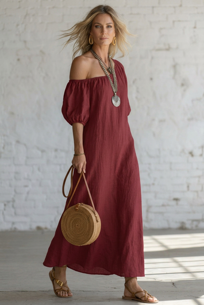 A woman wearing a flowing off-the-shoulder maxi dress styled with layered silver necklaces, gold hoop earrings, a woven straw tote bag, and strappy sandals, standing against a white brick wall.