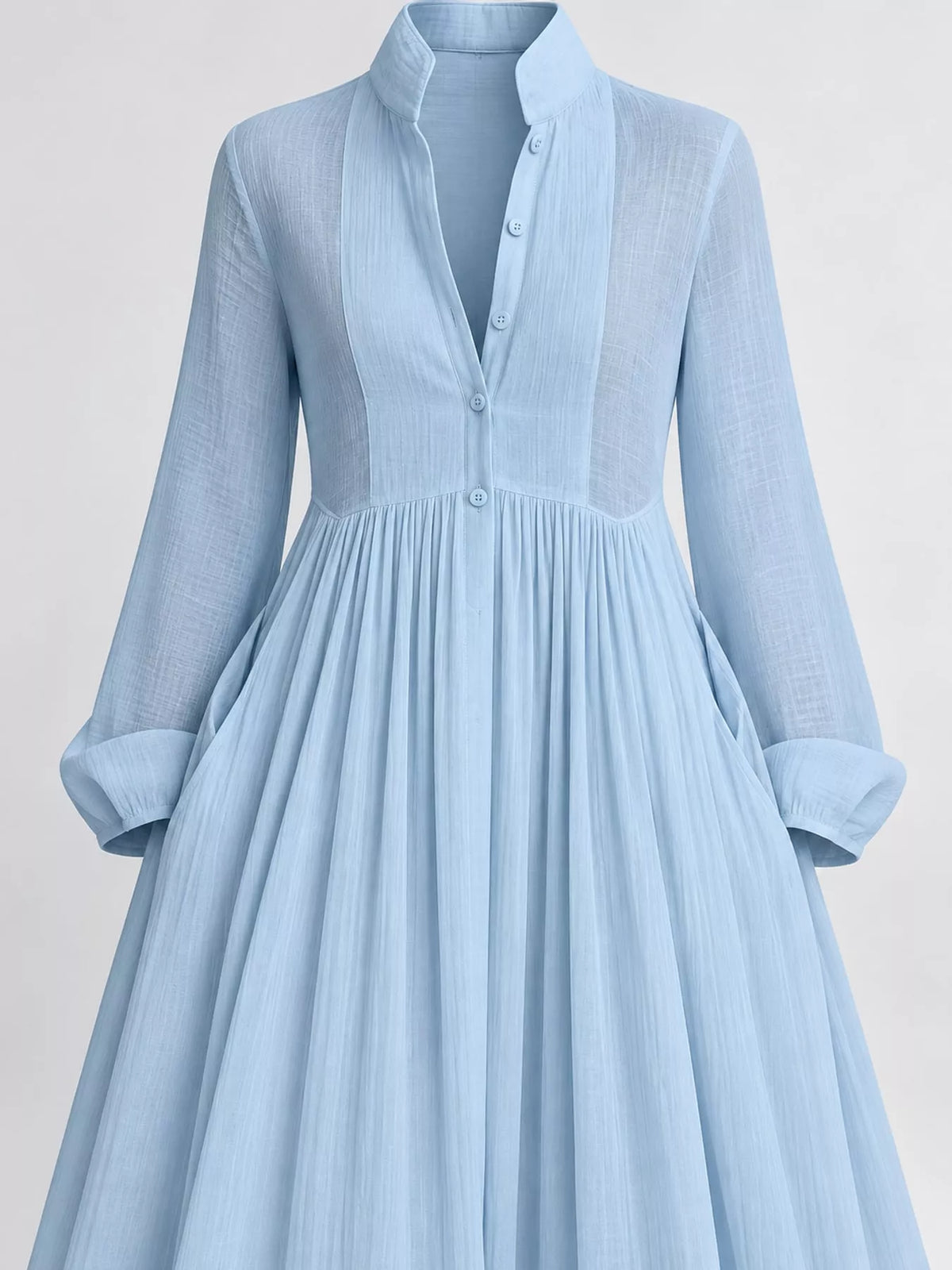 A_light_blue_long-sleeve_maxi_dress_with_a_deep_V-neckline_sheer_upper_chest_panel_and_side_pockets_standing_against_a_textured_gray_studio_backdrop.