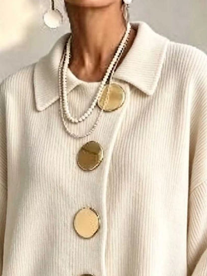 A close-up of a person wearing a cream ribbed knit collared sweater with oversized gold buttons, layered with pearl necklaces and dangling earrings.
