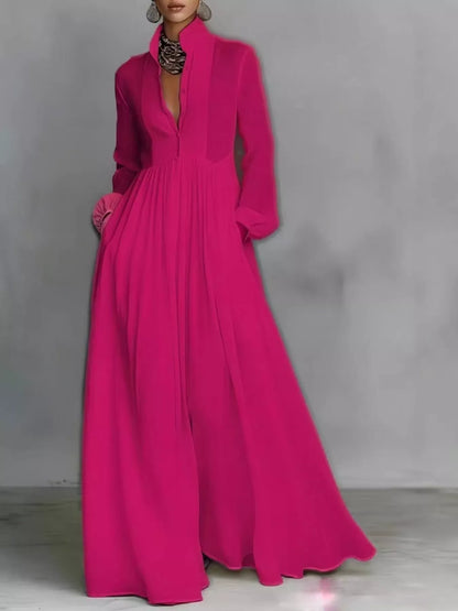 A woman models a long-sleeved, floor-length magenta maxi dress with a deep plunging V-neckline, high collar, and side pockets, set against a gray studio backdrop.
