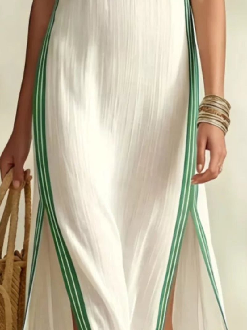 A sleeveless white maxi dress with green striped trim, worn by a model holding a straw bag in a beach-themed setting with palm leaves.