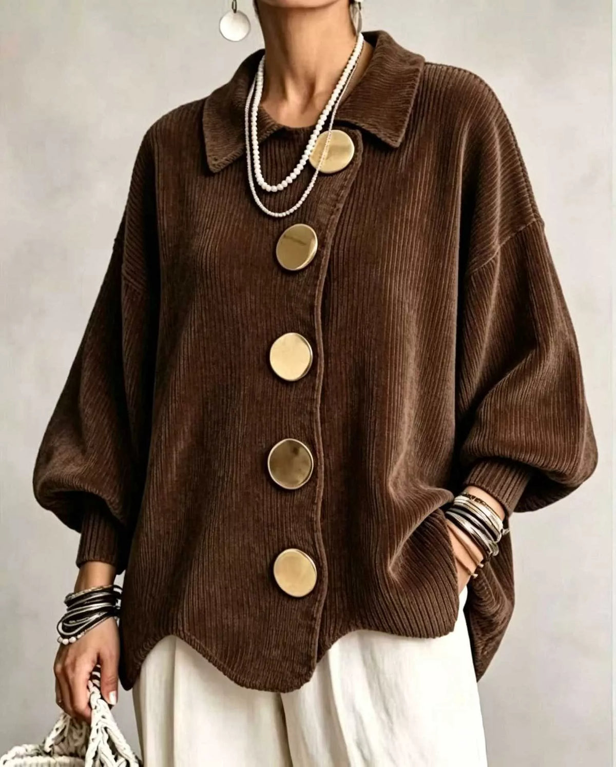 A woman wearing a brown corduroy button-front cardigan with oversized round gold-tone buttons, layered pearl necklaces, bracelets, and white pants, photographed against a soft neutral studio background.