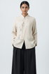A woman modeling a light beige linen long-sleeve top with a mandarin collar and delicate botanical embroidery on the front, paired with black wide-leg trousers, against a light gray studio background.