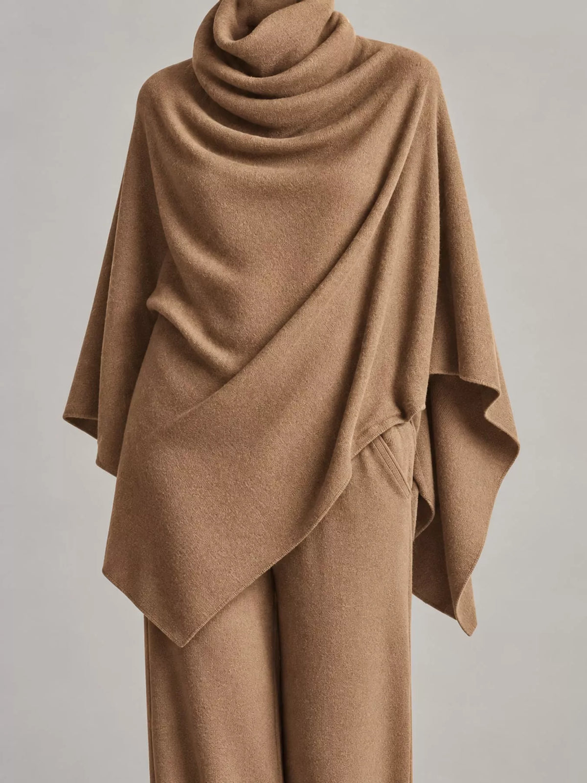 A camel brown draped knit poncho or shawl styled over matching wide-leg trousers, shown on a mannequin against a neutral light gray background.