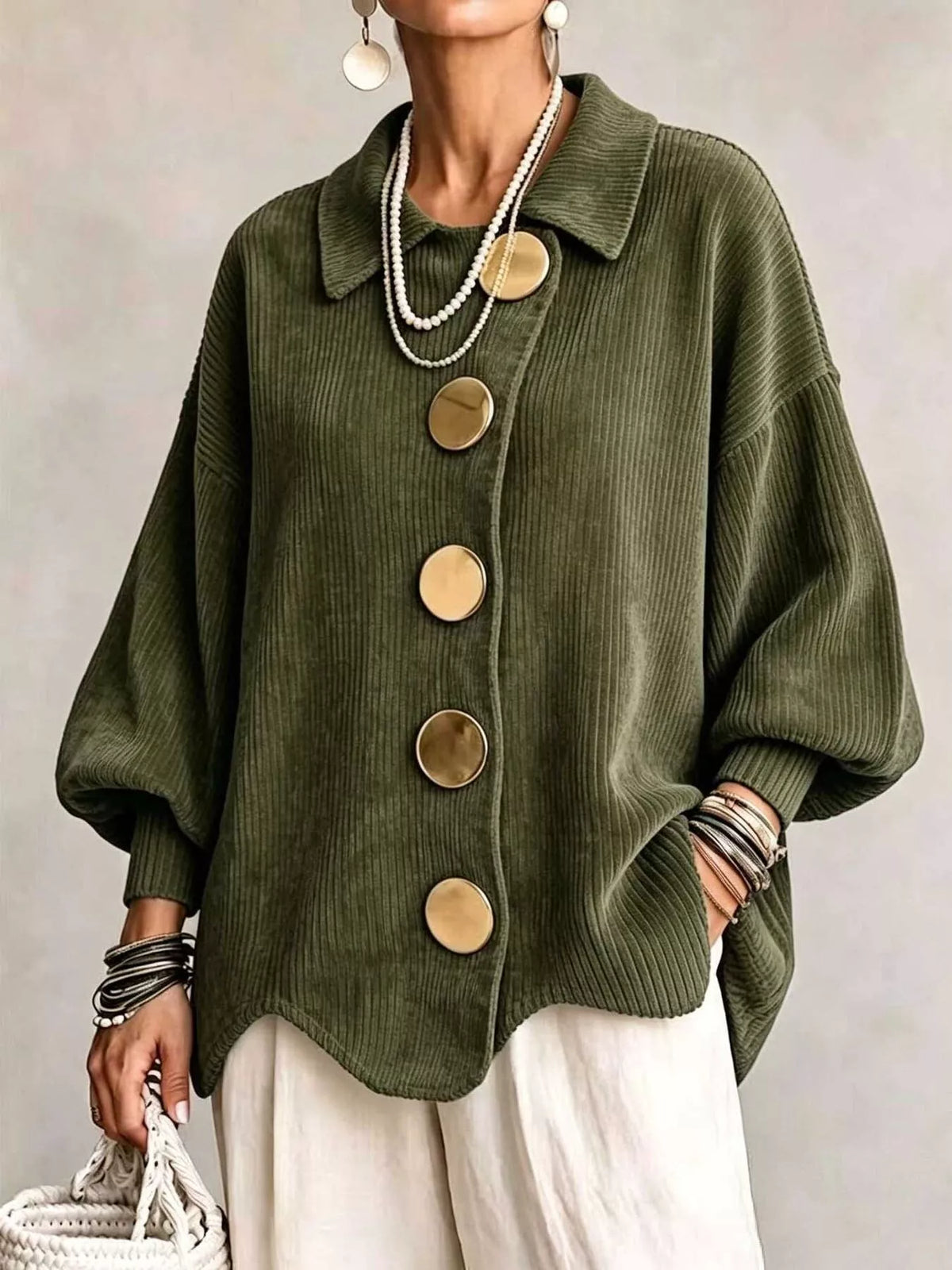 An olive green corduroy button-up cardigan with oversized round gold-tone buttons, worn by a model with white pants, layered pearl necklaces, and bracelets.