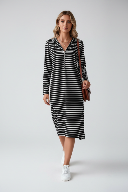 A woman modeling a black and white horizontally striped long-sleeve midi dress with a half-zip neckline, paired with white sneakers and a brown shoulder bag, against a light gray studio background.