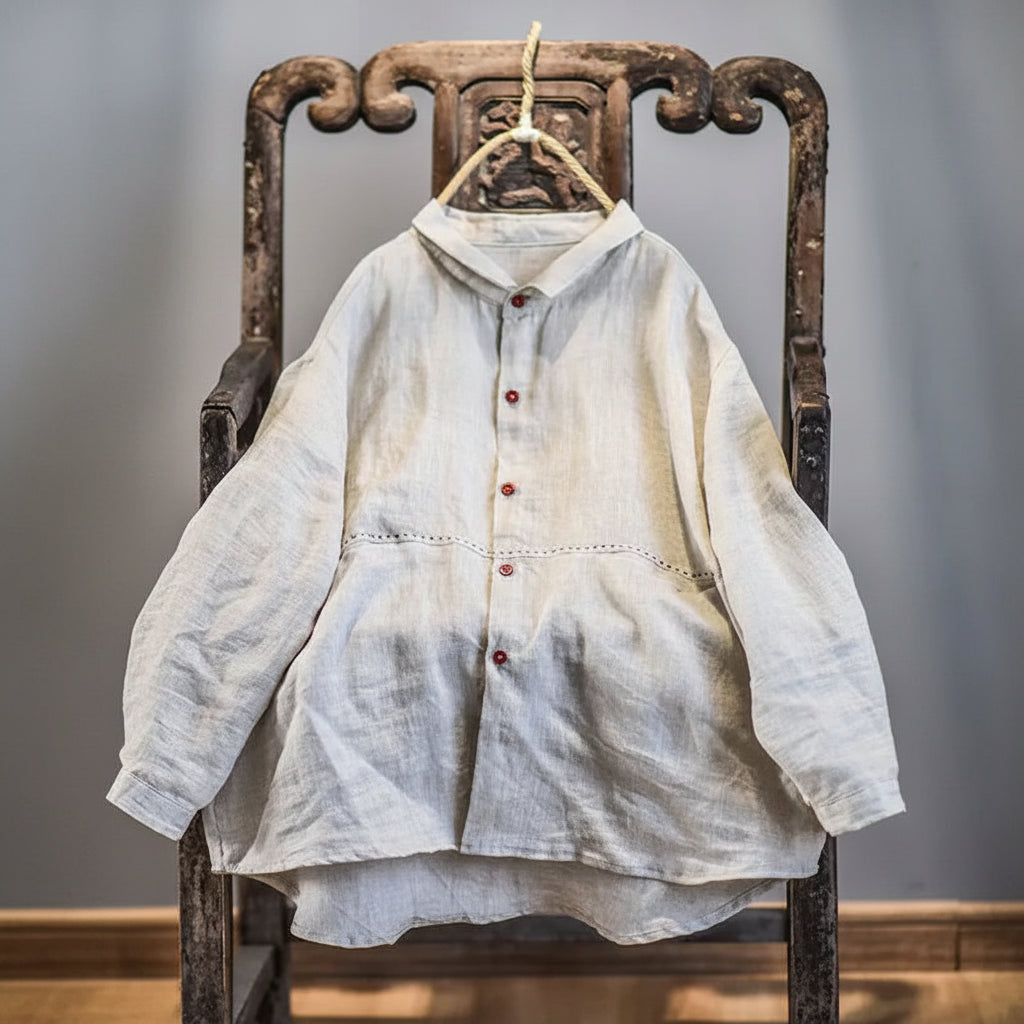 A light beige linen long-sleeve button-up shirt with red buttons, hanging on a rustic wooden chair.