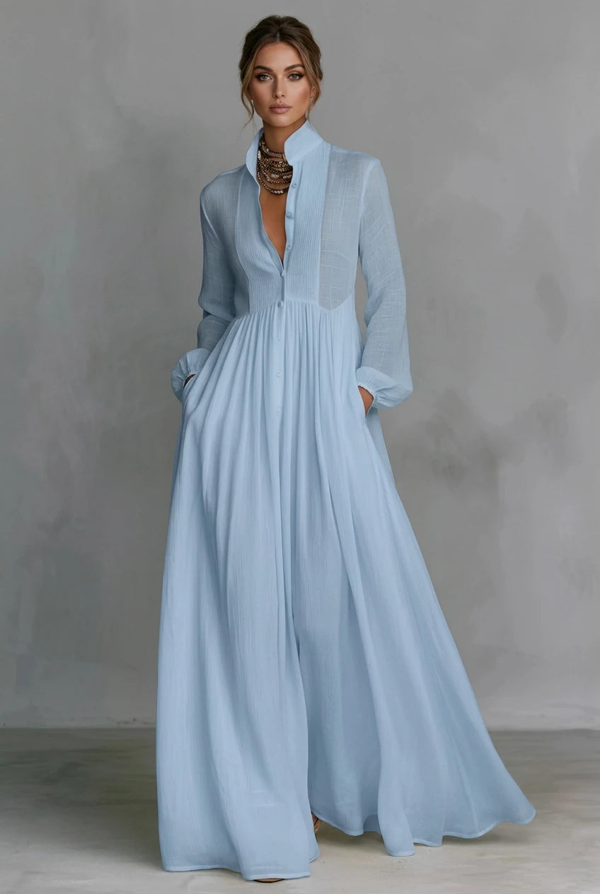 A_woman_wearing_a_light_blue_long-sleeve_maxi_dress_with_a_deep_V-neckline_sheer_upper_chest_panel_and_side_pockets_standing_against_a_textured_gray_studio_backdrop.