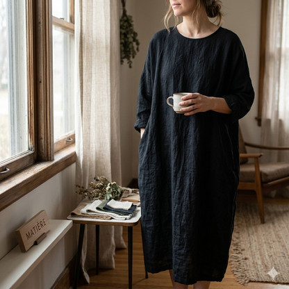 Woman in a dark blue dress holding a mug in a cozy room with a window and table.