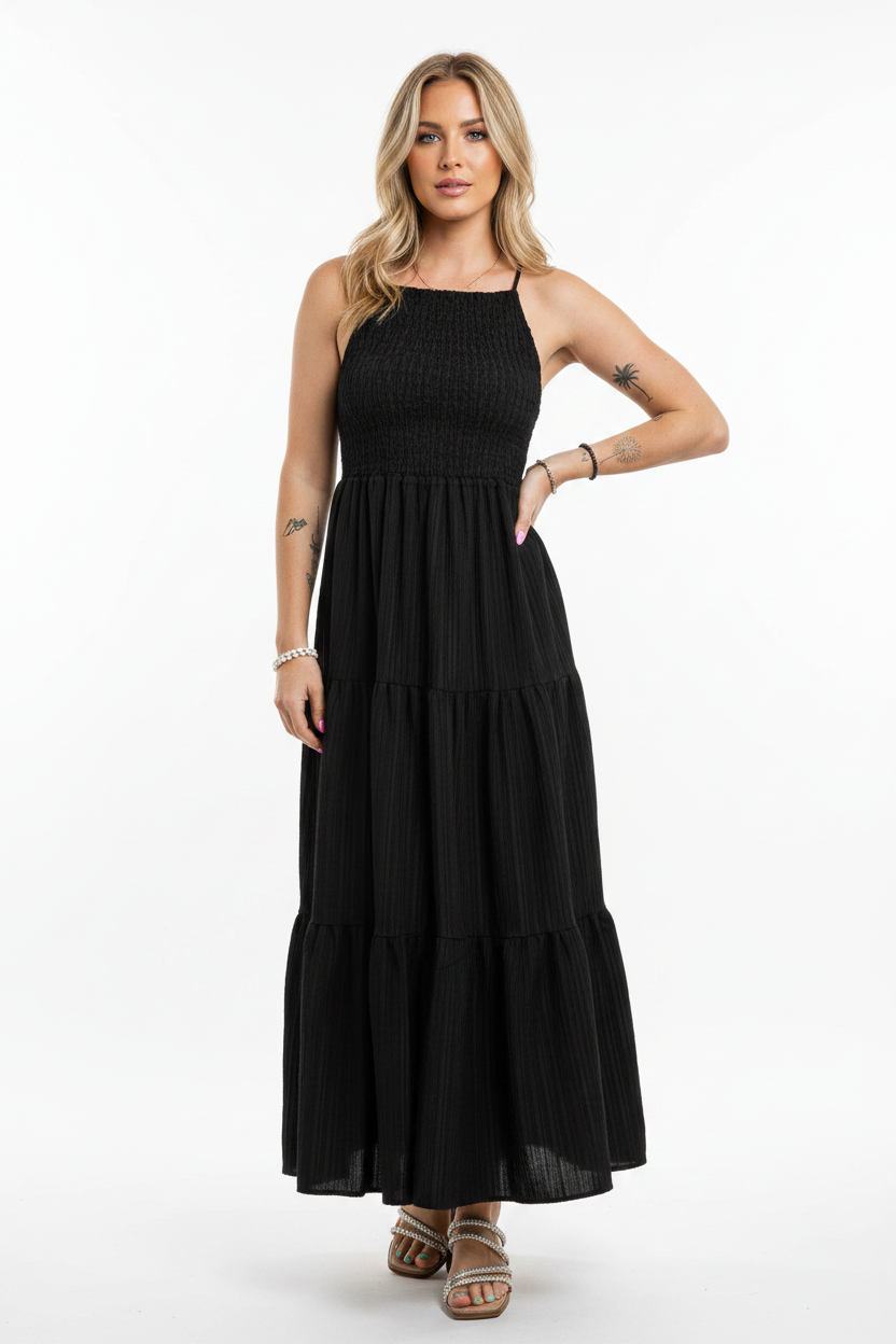 A woman modeling a black sleeveless maxi dress with a smocked bodice and tiered skirt, standing against a plain white studio background.