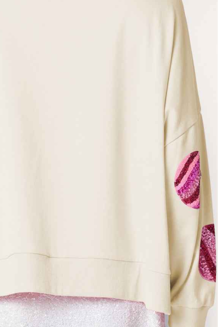 Close-up of a cream-colored sweatshirt with pink and purple sequined lip patch details on the sleeve, layered over a sparkly pink garment.