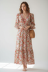 A woman stands barefoot in a bright studio wearing a long-sleeve floral maxi dress with a V-neck and gathered waist. She is holding a round woven straw bag.