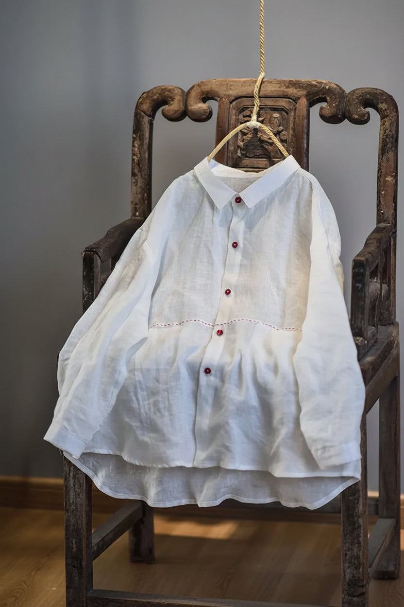 A white long-sleeve linen button-up shirt with red buttons, hanging on a wooden hanger from the back of a dark carved wooden chair.
